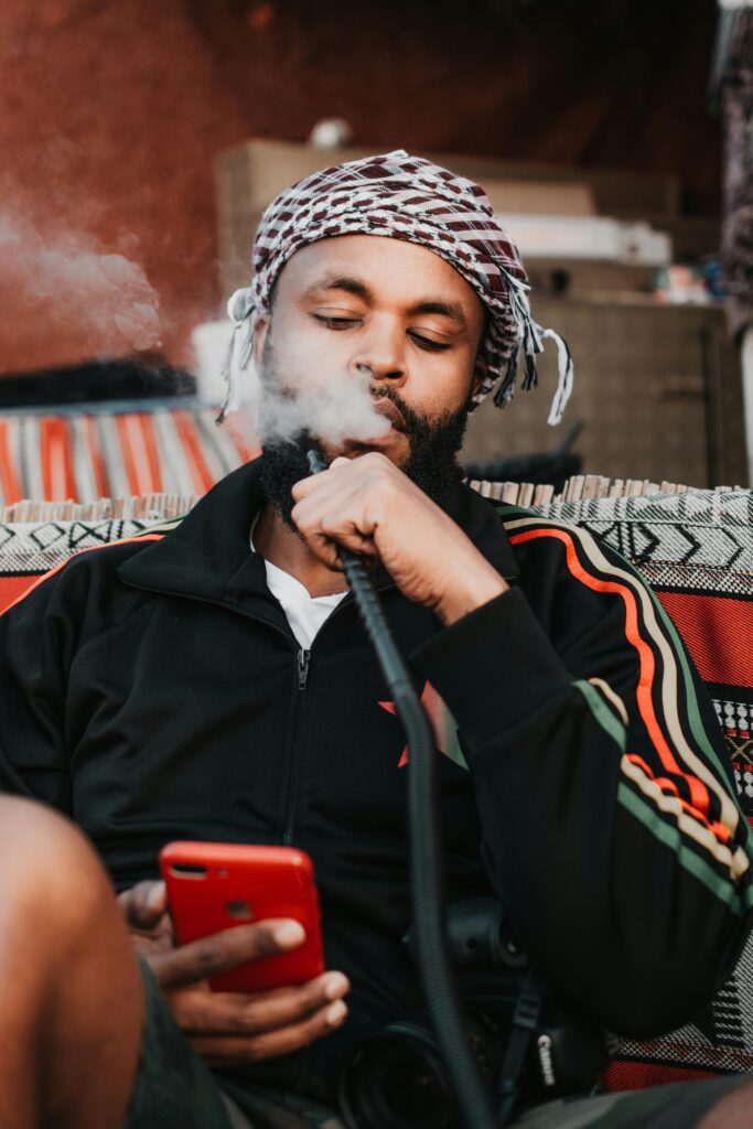 pexels-photo-3942712-3942712 Serious ethnic guy in casual clothes and traditional headdress browsing smartphone and smoking hookah in modern apartment